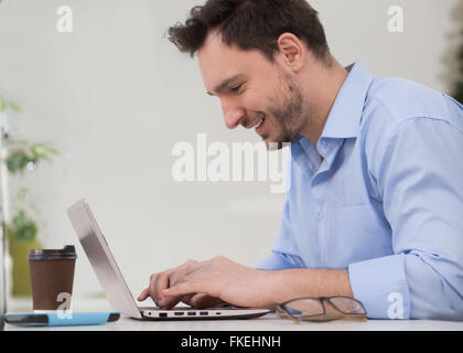 Close-up profile de bel homme à l'aide d'un ordinateur portable. Homme barbu en chemise bleu et souriant à l'ordinateur à l'écran. Banque D'Images