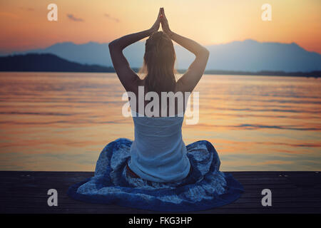 +Femme au coucher du soleil, heure de méditation en face d'un lac . Position de Yoga Banque D'Images