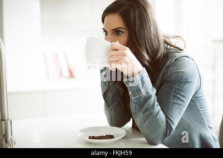 Jolie dame dans ses 30 ans de boire du café dans sa cuisine portant chemise en denim bleu sur son teabreak Banque D'Images