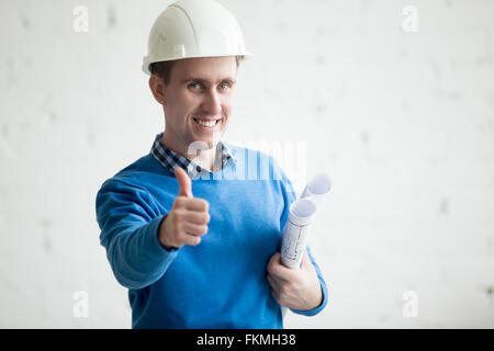 Portrait of young cheerful handsome ingénieur dans white hard hat holding plans du projet architectural en agence. Friendly Banque D'Images
