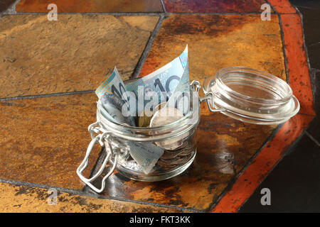 Un petit contenant en verre utilisé pour économiser de l'argent australien, à la fois des billets et des pièces de monnaie, placé sur une table carrelée Banque D'Images