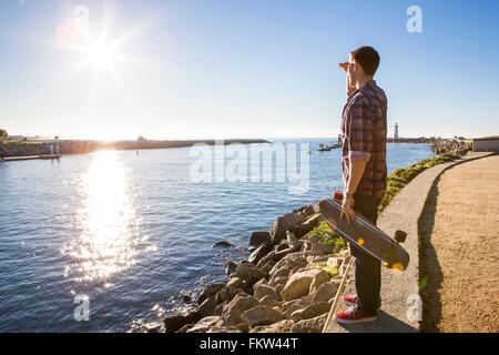 Jeune homme, holding skateboard, debout au bord de l'eau, looking at view Banque D'Images