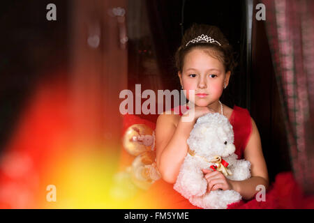 Une adorable petite fille, habillée d'une robe rouge luxuriant, serrant un ours en peluche à côté de la fenêtre. Arrière-plan flou Banque D'Images