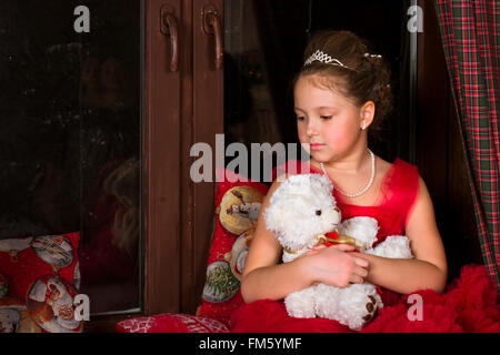 Une adorable petite fille, habillée d'une robe rouge luxuriant, serrant un ours en peluche à côté de la fenêtre Banque D'Images