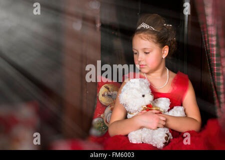 Une adorable petite fille, habillée d'une robe rouge luxuriant, serrant un ours en peluche à côté de la fenêtre Banque D'Images