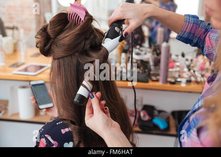 Coiffure femme coiffure rendant l'utilisation de fer à friser pour cheveux longs de jeunes femmes avec le smartphone en instituts de beauté Banque D'Images
