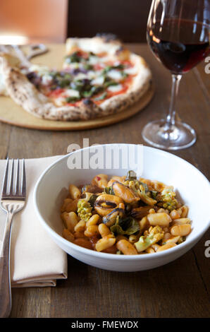 Un bol de haricot blanc, romanesco et les moules soupe avec du vin rouge et une pizza sur la table derrière elle.