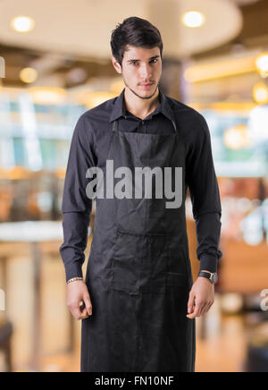 Jeune cuisinier ou serveur posant, le port de tablier noir et chemise en bar ou restaurant ou dîner Banque D'Images