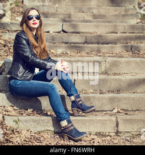 Jeune femme élégante en jeans et blouson de cuir reposant sur l'escalier du parc. Jolie jeune fille aux cheveux longs sitting on steps Banque D'Images