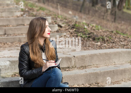 Jeune femme assise sur les marches dans le parc des dessins d'un pad. Thoughtful woman avec ordinateur portable sur des marches en pierre Banque D'Images