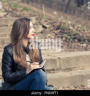 Jeune femme assise sur les marches dans le parc l'écriture dans un tampon. Jolie fille adultes faire des croquis dans le bloc-notes sur les mesures Banque D'Images