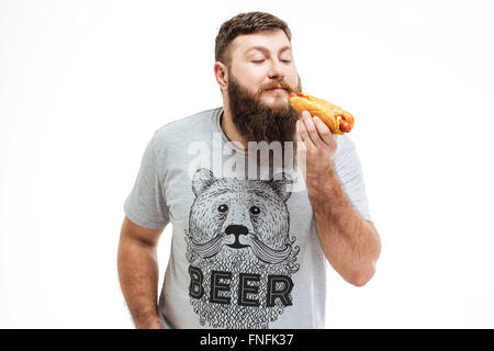 Heureux homme barbu debout et eating hot dog sur fond blanc Banque D'Images