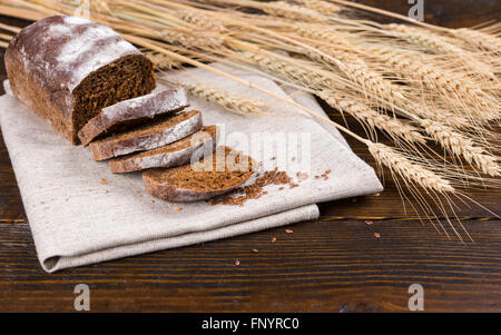 Tiges de tiges de blé entier et de grains en regard de simple miche de pain de seigle délicieux avec des tranches de tissu brun sur table en bois sombre. Banque D'Images