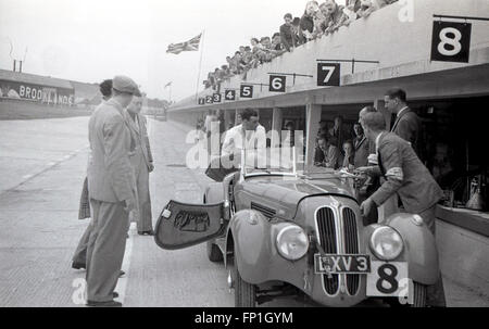 Années 1930, photo historique de la voie des stands du circuit de Brooklands, le premier circuit de course automobile au monde construit à cet effet, Weybridge, Surrey, Angleterre. Une voiture de course monoplace à toit ouvert, no 8, garée dans une voie sur le côté de la piste avec des spectateurs assis au-dessus du sol regardant. Banque D'Images