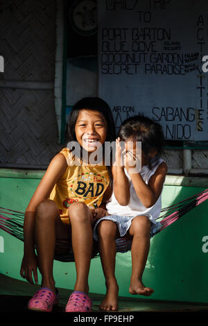 Deux filles assis au soleil dans un hamac pendu sur le mur en face d'un magasin local sourire, rire, se cache un peu Banque D'Images