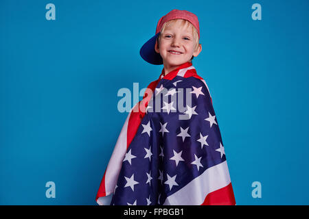 Smiling boy enveloppée de drapeau américain isolé sur fond bleu Banque D'Images