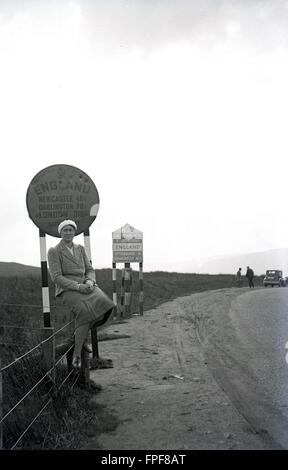 Historique années 1930, dame assise sur une clôture en bordure de la route en face de l'AA signe pour l'Angleterre - Newcastle, Darlington et Londres (318 miles) 1/4 - à la frontière avec l'Écosse. Banque D'Images