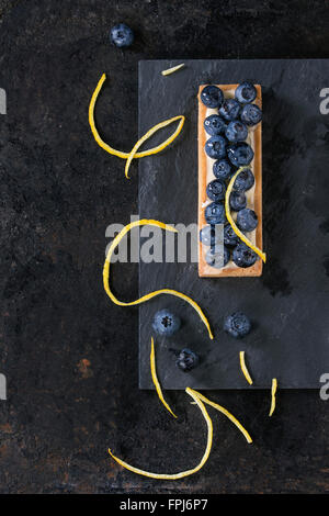 Tartelette au citron carrés avec des bleuets frais, servi sur ardoise noire avec les zestes de citron sur fond noir. Mise à plat. Avec s Banque D'Images