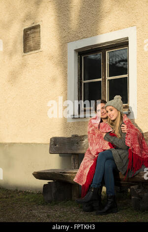 Jeune couple enveloppé dans une couverture assis sur un banc pendant le coucher du soleil, Munich, Bavière, Allemagne Banque D'Images