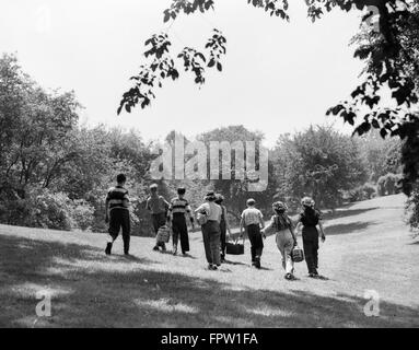 1950 GROUPE DE HUIT ADOLESCENTS Garçons Filles MARCHER EN ÉTÉ CHAMP SOLEIL PORTANT DES PANIERS DE PIQUE-NIQUE Banque D'Images