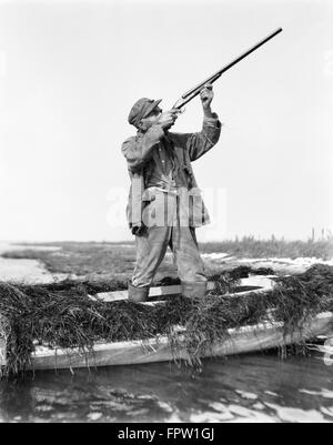 Années 1920 L'HOMME LA CHASSE AUX CANARDS DEBOUT DANS SNEAKBOX BATEAU DANS LA BAIE DE BARNEGAT FUSIL MARAIS VISANT NEW JERSEY USA Banque D'Images