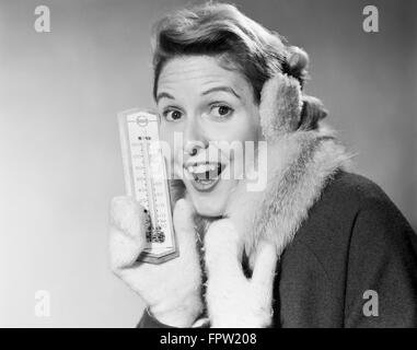 1950 SMILING WOMAN LOOKING AT CAMERA EN HIVER FROID THERMOMÈTRE HOLDING portant des mitaines MANTEAU CASQUE ANTIBRUIT Banque D'Images