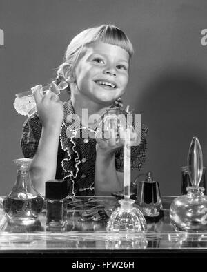 1960 SMILING LITTLE GIRL SITTING AT MOTHER'S BUREAU VANITÉ DES FLACONS DE PARFUM EN FACE À LA CAMÉRA EN MIROIR DANS Banque D'Images