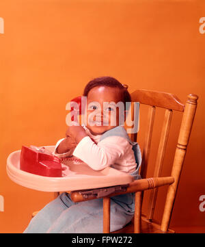 1970 AFRICAN AMERICAN BABY SITTING CHAISE HAUTE BÉBÉ HOLDING TOY ROUGE Téléphone Banque D'Images