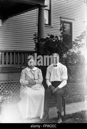 Années 1900 PORTRAIT HOMME FEMME COUPLE ASSIS SUR DES CHAISES EN FACE DE HOUSE LOOKING AT CAMERA Banque D'Images