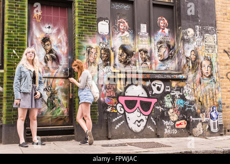 Deux filles de taille basse se tenant devant un mur recouvert de graffiti et de peintures murales représentant les visages de personnages célèbres du cinéma Banque D'Images