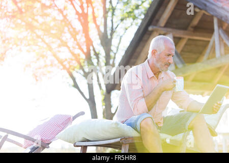 Senior man drinking coffee and using digital tablet on lounge chair in backyard Banque D'Images