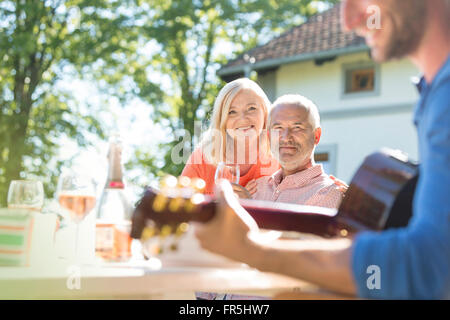 Senior couple drinking wine et regarder des fils adultes à jouer de la guitare sur un patio ensoleillé Banque D'Images