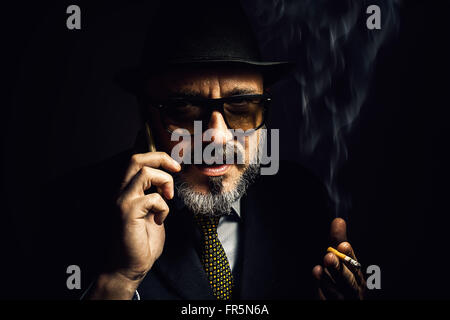 Portrait d'un homme plus âgé, portant un chapeau et veste classique, fumer un cigare et de parler par téléphone. Banque D'Images