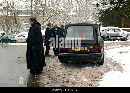 Un prêtre dans une longue cape noire attend dans la neige par un corbillard à la suite d'un enterrement au pays de Galles Banque D'Images