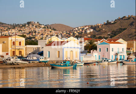 Pedi, Symi, sud de la mer Egée, en Grèce. Les bâtiments au bord de l'eau éclairée par le soleil levant, le village perché de Horio en arrière-plan. Banque D'Images