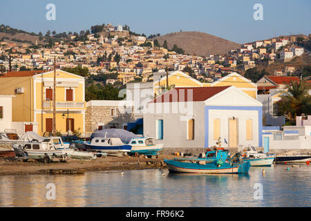 Pedi, Symi, sud de la mer Egée, en Grèce. Les bâtiments au bord de l'eau éclairée par le soleil levant, le village perché de Horio en arrière-plan. Banque D'Images