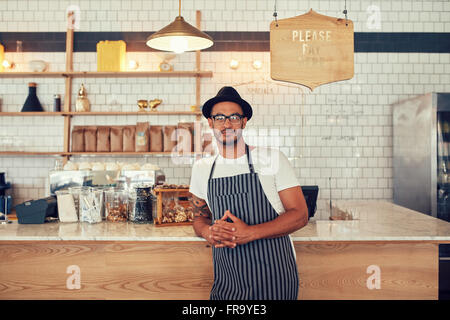 Portrait of a confident young coffee shop owner standing at the cafe counter. Handsome young man working at a restaurant and wea Banque D'Images
