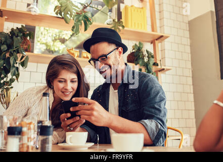 Portrait of young friends sitting in cafe à la recherche aux photos sur téléphone mobile. Heureux les jeunes à une réunion dans un restaurant usin Banque D'Images