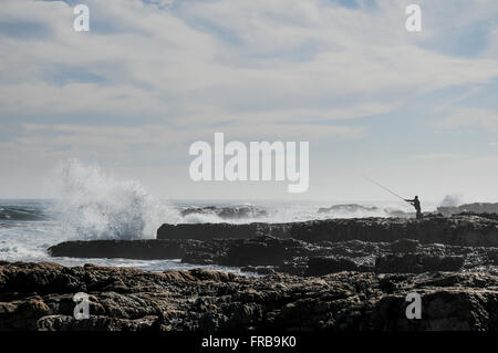 Un homme à la pêche la plus au sud de la ville du Cap point jette sa canne à pêche comme des vagues s'écraser sur les rochers. Banque D'Images