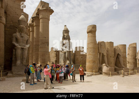 Les groupes de touristes se lever et photographie sous les colosses géant dans la cour de Ramsès II, à l'Egypte antique du temple de Louxor, vallée du Nil, en Égypte. Le temple a été construit par Amenhotep III, complété par Toutânkhamon puis ajouté à par Ramsès II. Vers l'arrière est un granit sanctuaire dédié à Alexandre le Grand et dans une autre partie, était un camp romain. Le temple a été presque continue à utiliser comme lieu de culte jusqu'à nos jours. Banque D'Images