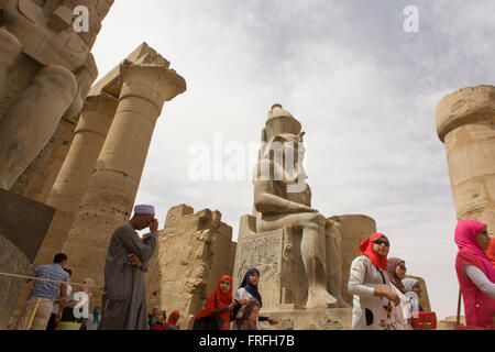 Les groupes de touristes se lever et photographie sous le colosse géant dans la cour de Ramsès II, à l'Egypte antique du temple de Louxor, vallée du Nil, en Égypte. Le temple a été construit par Amenhotep III, complété par Toutânkhamon puis ajouté à par Ramsès II. Vers l'arrière est un granit sanctuaire dédié à Alexandre le Grand et dans une autre partie, était un camp romain. Le temple a été presque continue à utiliser comme lieu de culte jusqu'à nos jours. Banque D'Images