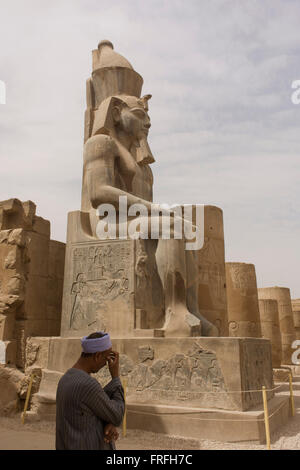 Un guide local et gardien sous le colosse stitting massive dans la cour de Ramsès II à l'Egypte antique du temple de Louxor, vallée du Nil, en Égypte. Le temple a été construit par Amenhotep III, complété par Toutânkhamon puis ajouté à par Ramsès II. Vers l'arrière est un granit sanctuaire dédié à Alexandre le Grand et dans une autre partie, était un camp romain. Le temple a été presque continue à utiliser comme lieu de culte jusqu'à nos jours. Banque D'Images