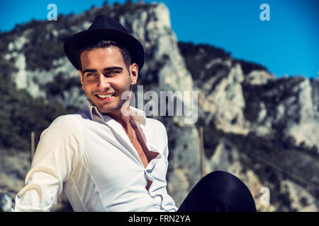 Les jeunes bel homme élégant en chemise blanche et chapeau fedora noir, sur la plage en regardant loin de sourire. Sur fond de collines Banque D'Images