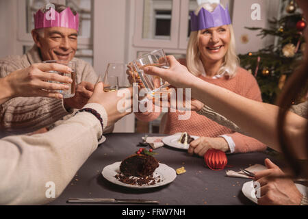 Happy senior couple with paper crowns clinking glasses tout en ayant le pudding de Noël avec leur famille Banque D'Images