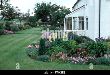 Géraniums vivaces bleu et jaune pâle avec lupin dianthus rose dans une frontière d'été à côté d'une petite maison blanche Banque D'Images