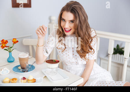 Cheerful pretty young woman avec de beaux cheveux bouclés dessert manger et boire un café latte dans Banque D'Images