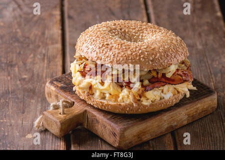 Bagel avec bacon, œufs brouillés et oignon frit sur petite planche à découper en bois sur fond de bois. Banque D'Images