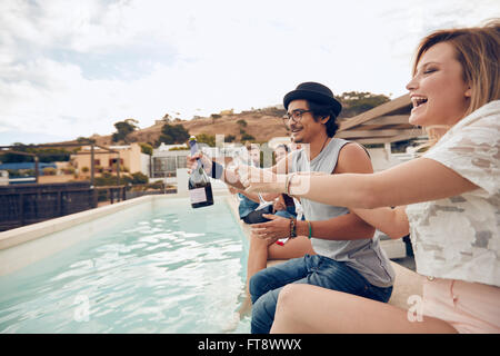 Tourné en extérieur de professionnels jeunes assis sur le bord de la piscine de boire du champagne. Friends toasting et multiraciale Banque D'Images