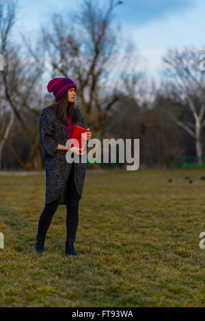 Un adolescent girl in purple hat holding livre avec couvercle rouge en étant debout sur une pelouse dans un parc et à la voiture. Banque D'Images