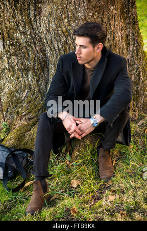 Handsome young man leaning against tree, looking at camera, dans une journée ensoleillée portant un manteau noir Banque D'Images